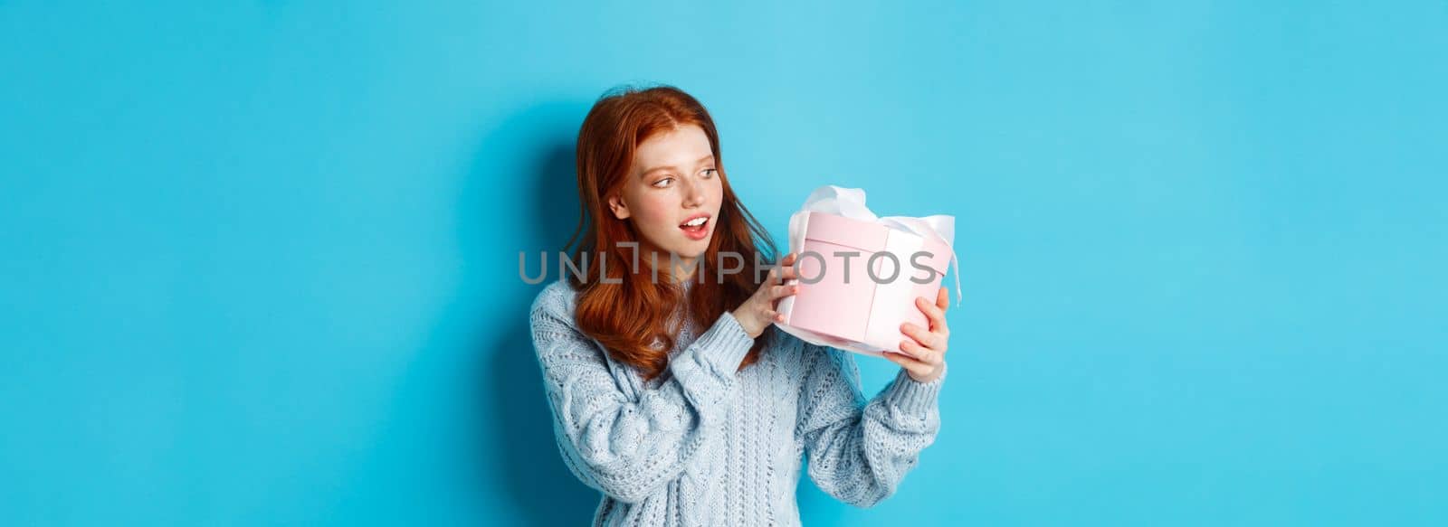 Curious teen girl with red hair, shaking gift box and wonder what inside, standing over blue background by Benzoix