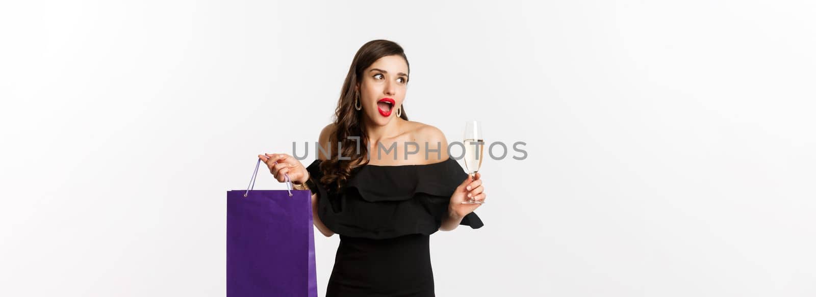 Excited woman shop and drink champagne, holding shopping bag, looking amazed, standing against white background by Benzoix