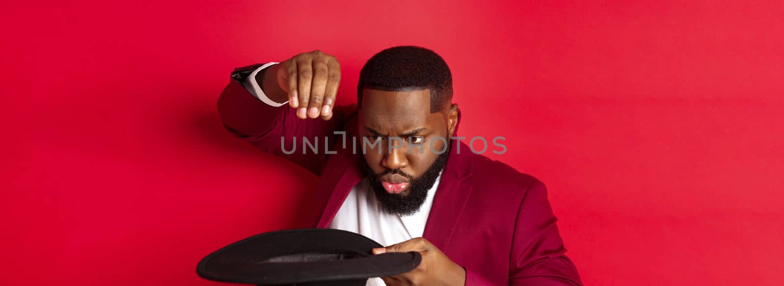 Close-up of charismatic Black male magician perform a trick with his hat, performing on christmas party, standing over red background.