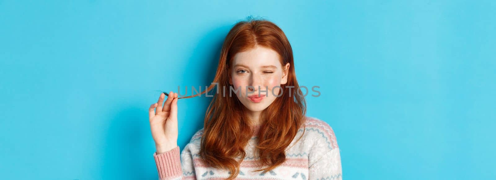 Close-up of cheeky redhead girl playing with hair strand, winking and smiling at camera, standing over blue background.