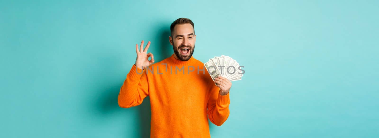 Shopping. Excited guy holding money, showing ok sign and winking, standing over light blue background by Benzoix
