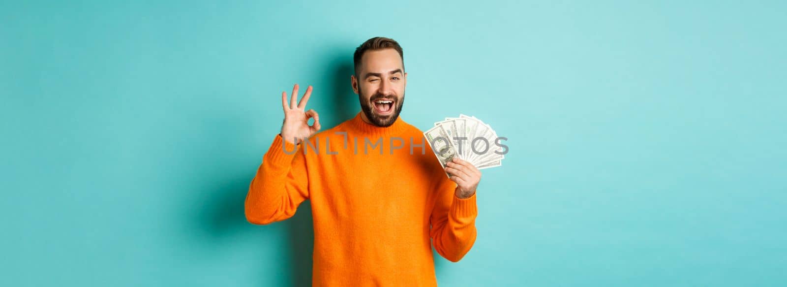 Shopping. Excited guy holding money, showing ok sign and winking, standing over light blue background. Copy space