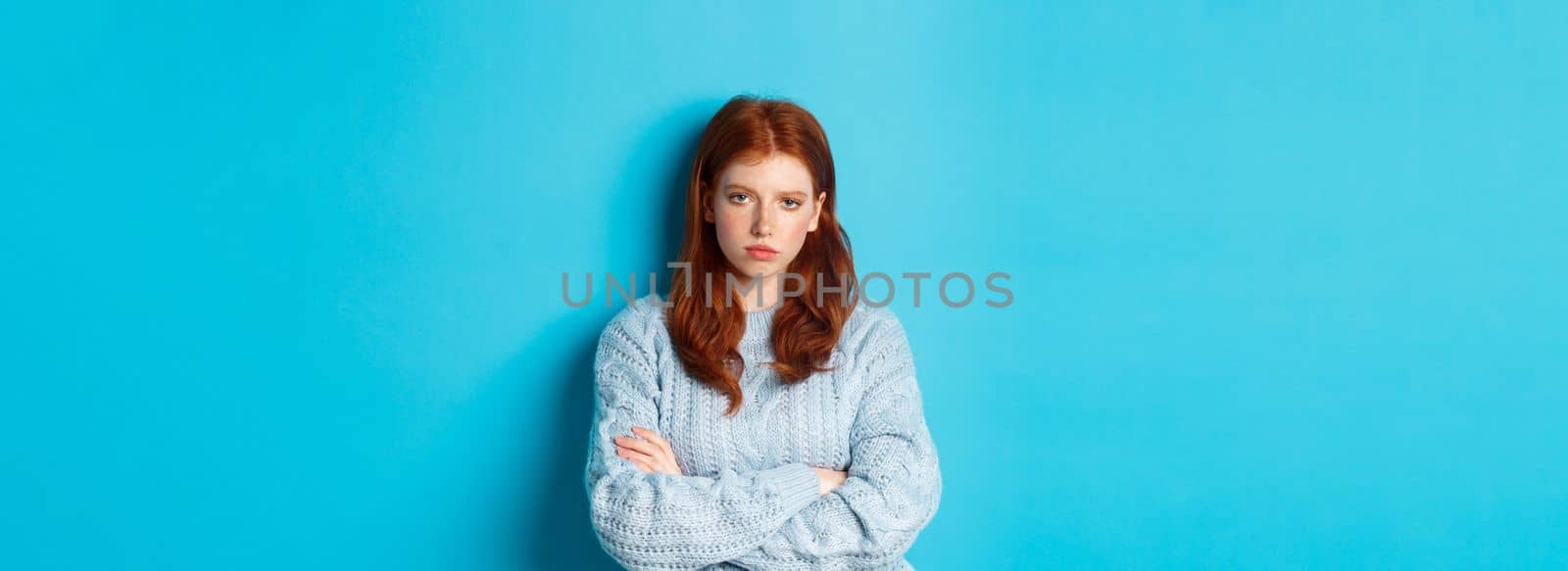 Annoyed and bothered redhead teeange girl cross arms on chest, staring at something lame and boring, standing against blue background.