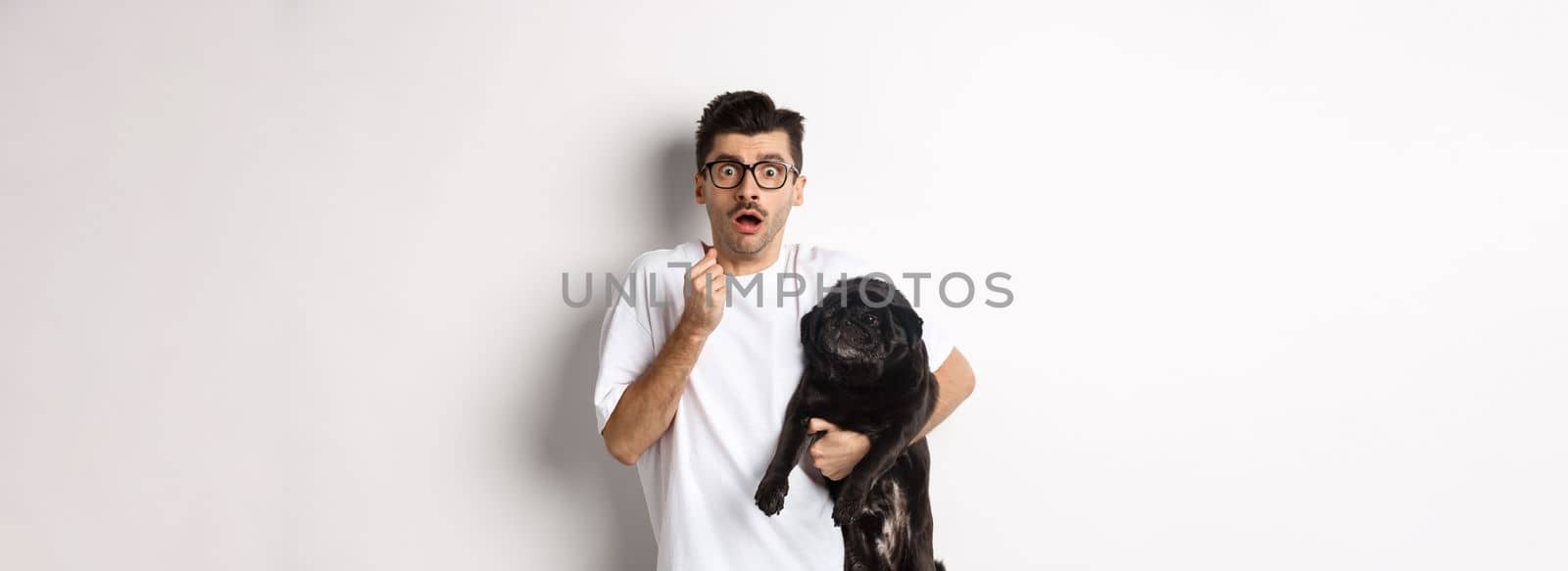 Scared hipster hugging his cute black puppy and staring at camera frightened. Dog owner looking shocked, holding pug, standing over white background by Benzoix