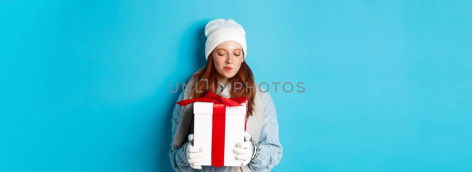 Winter holidays and Christmas sales concept. Intrigued redhead girl holding present, curiously staring at box with gift, trying guess what inside, standing over blue background.