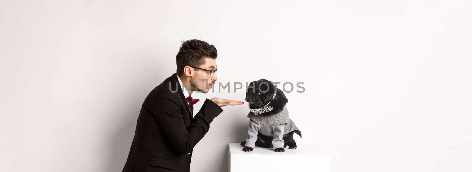 Pets, winter holidays and celebration concept. Handsome young man sending air kiss at cute black puppy wearing costume for New Year, owner standing in suit over white background by Benzoix