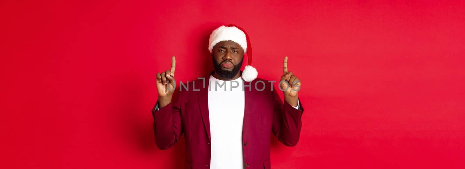 Christmas, party and holidays concept. Miserable and sad african american man pointing fingers up, looking disappointed, wearing santa hat, red background by Benzoix