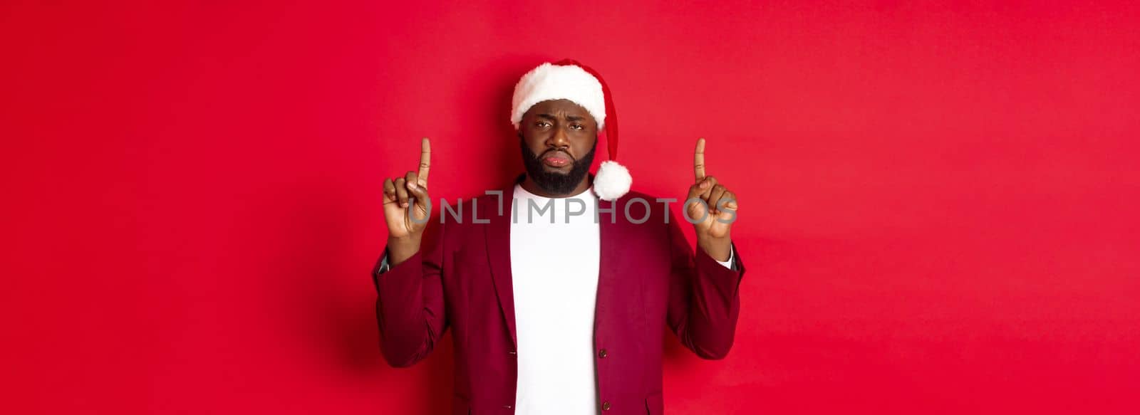 Christmas, party and holidays concept. Miserable and sad african american man pointing fingers up, looking disappointed, wearing santa hat, red background.