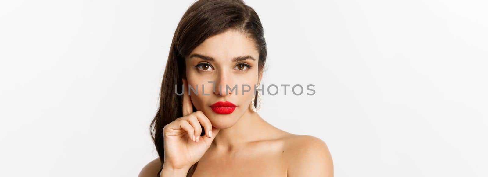 Fashion and beauty concept. Close-up of elegant beautiful woman in black dress, evening makeup and red lipstick, looking sassy at camera, standing over white background.