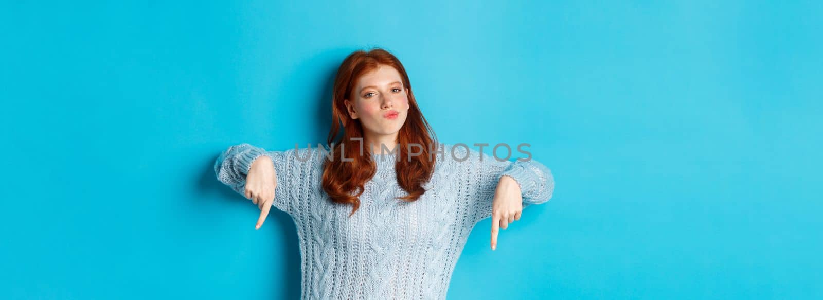 Winter holidays and people concept. Thoughtful teenage redhead girl in sweater, pointing fingers down and pondering, making decision, standing over blue background.