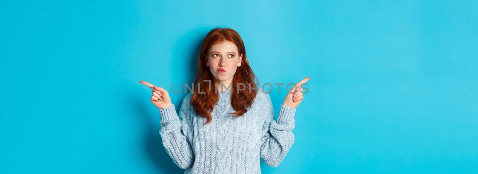 Winter holidays and people concept. Thoughtful redhead girl making decision, pointing fingers sideways, choosing between two ways, standing over blue background by Benzoix