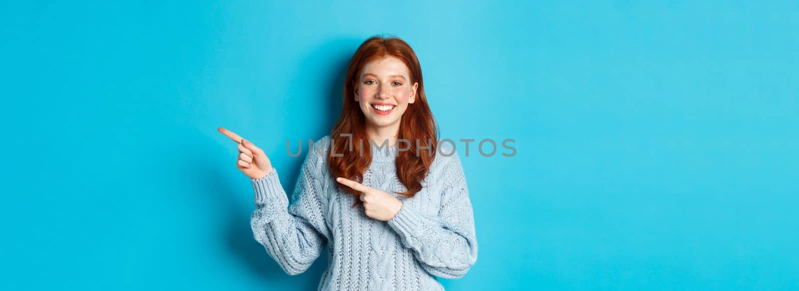 Winter holidays and people concept. Pretty teenage girl with red hair, pointing fingers right at logo copy space, smiling at camera, blue background.