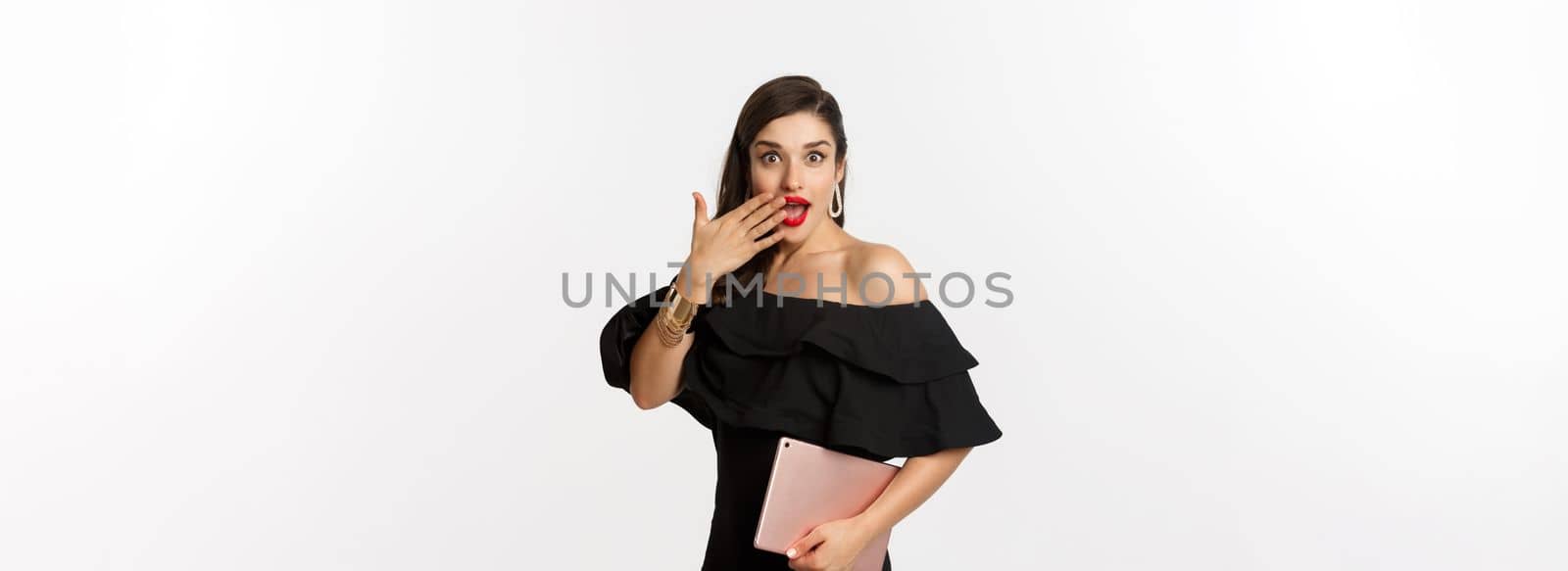 Fashion and shopping concept. Stylish young woman with glamour makeup, wearing black dress, holding digital tablet and looking surprised, white background.