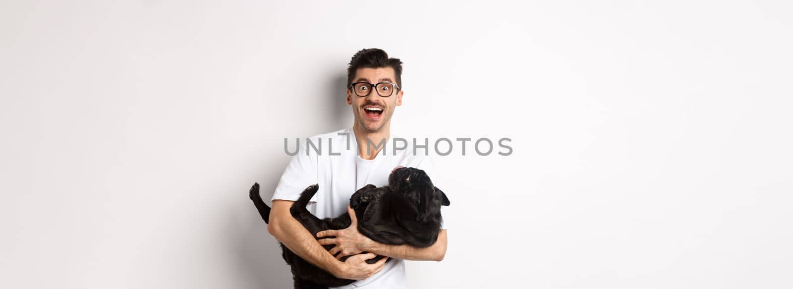 Handsome young man playing with cute black puppy. Dog owner petting a pug, standing over white background.