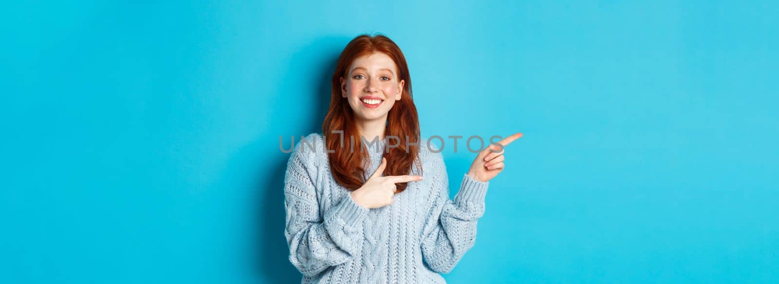 Cute teenager girl with red hair and freckles, pointing fingers left at logo and smiling, showing advertisement, standing over blue background.