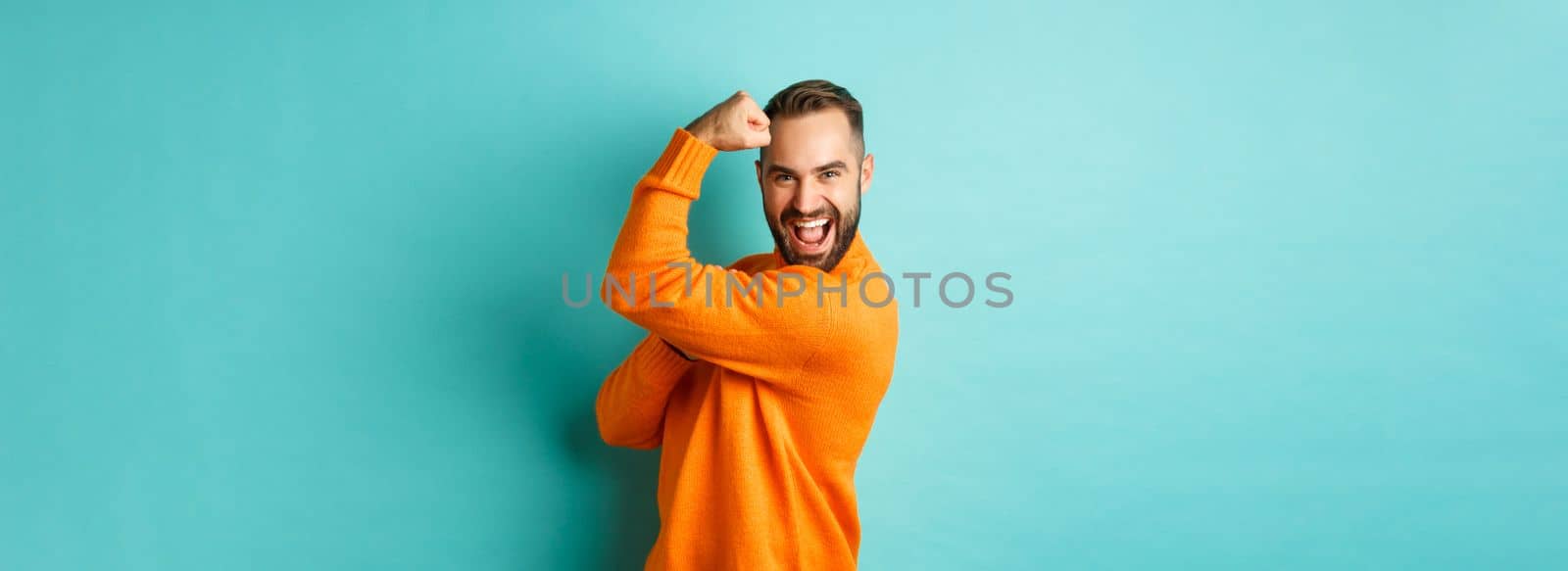 Confident handsome man show-off his muscles, flex biceps, feeling strong, standing over light blue background.