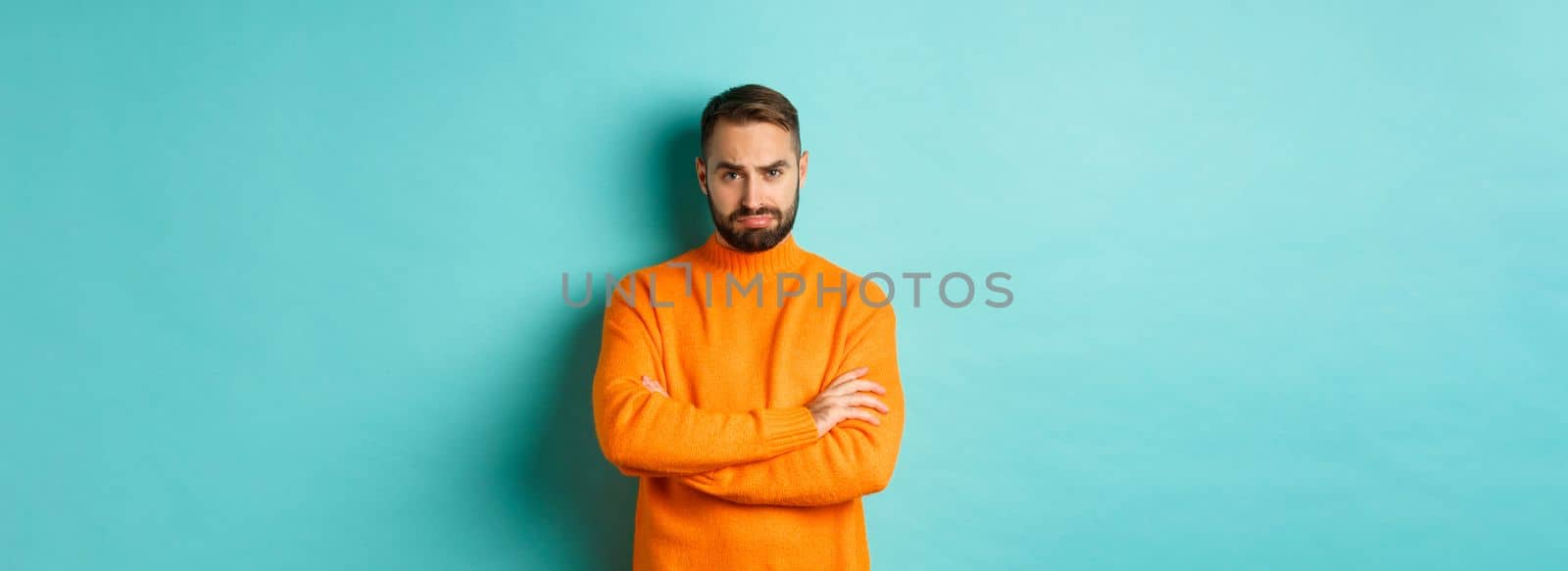 Silly sad guy with beard, sulking and looking offended, cross arms on chest and frowning upset, standing over light blue background.