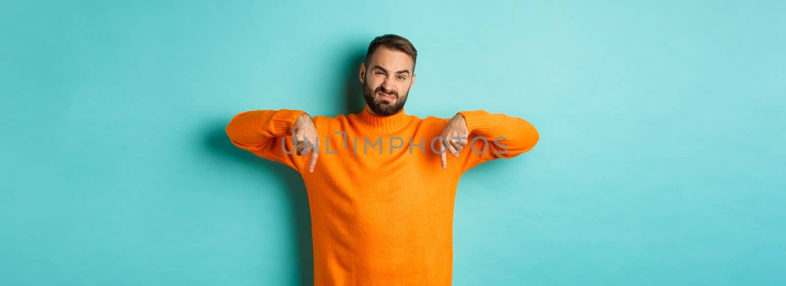Displeased and skeptical handsome man pointing fingers down, showing bad product, dislike store or advertisement, grimacing disappointed, standing over turquoise background by Benzoix
