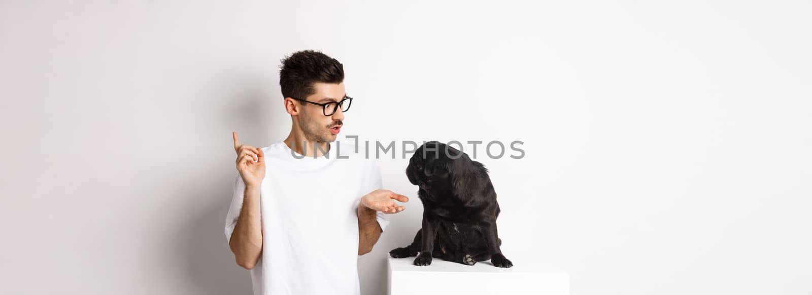 Handsome young man teaching dog commands, talking to cute black pug, standing over white background by Benzoix