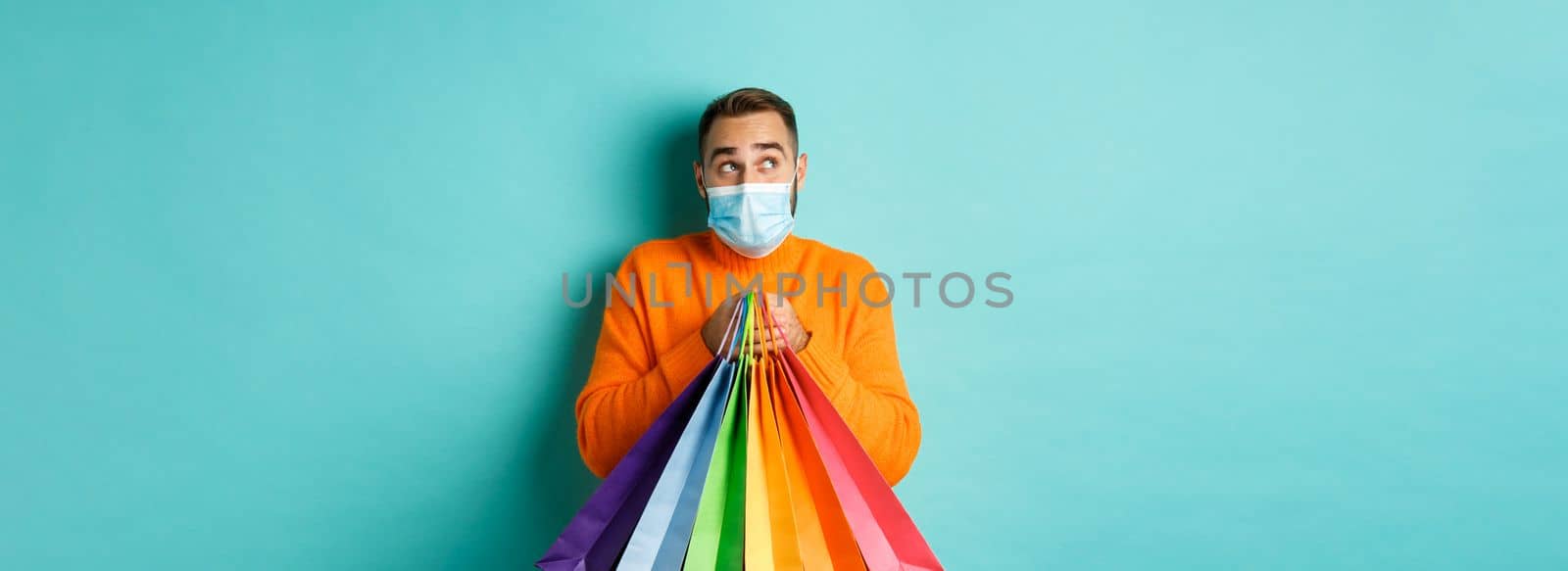 Covid-19, pandemic and lifestyle concept. Man thinking, wearing face mask, holding shopping bags, imaging something, standing over turquoise background by Benzoix