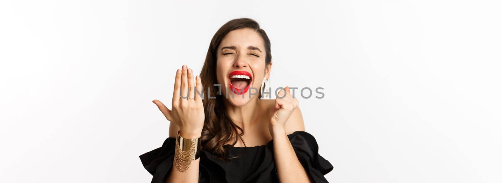 Fashion and beauty concept. Excited and happy woman said yes, showing hand with engagement ring and shouting joyful, become bride, standing over white background by Benzoix