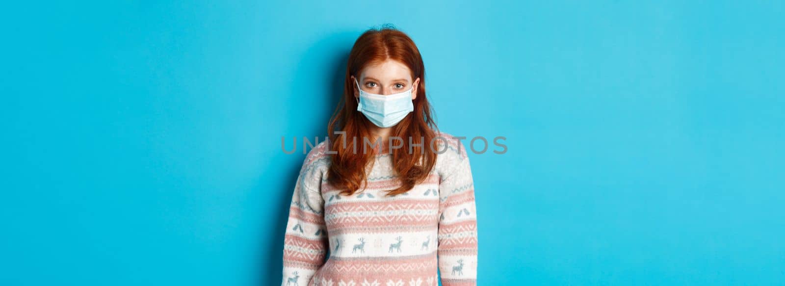 Winter, covid-19 and quarantine concept. Young redhead girl in sweater and face mask staring at camera, standing over blue background by Benzoix