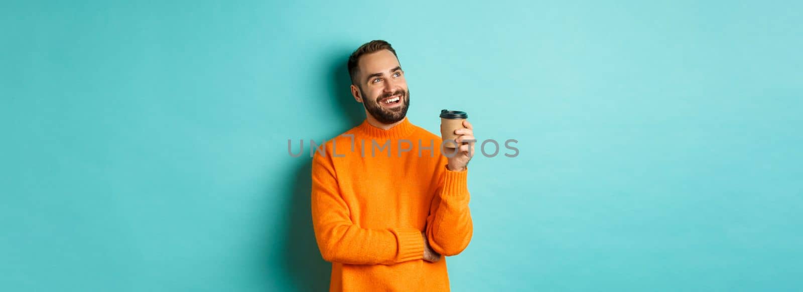 Thoughtful smiling man looking at upper left corner, having idea while drinking coffee from takeaway cup, standing in orange sweater against turquoise background.