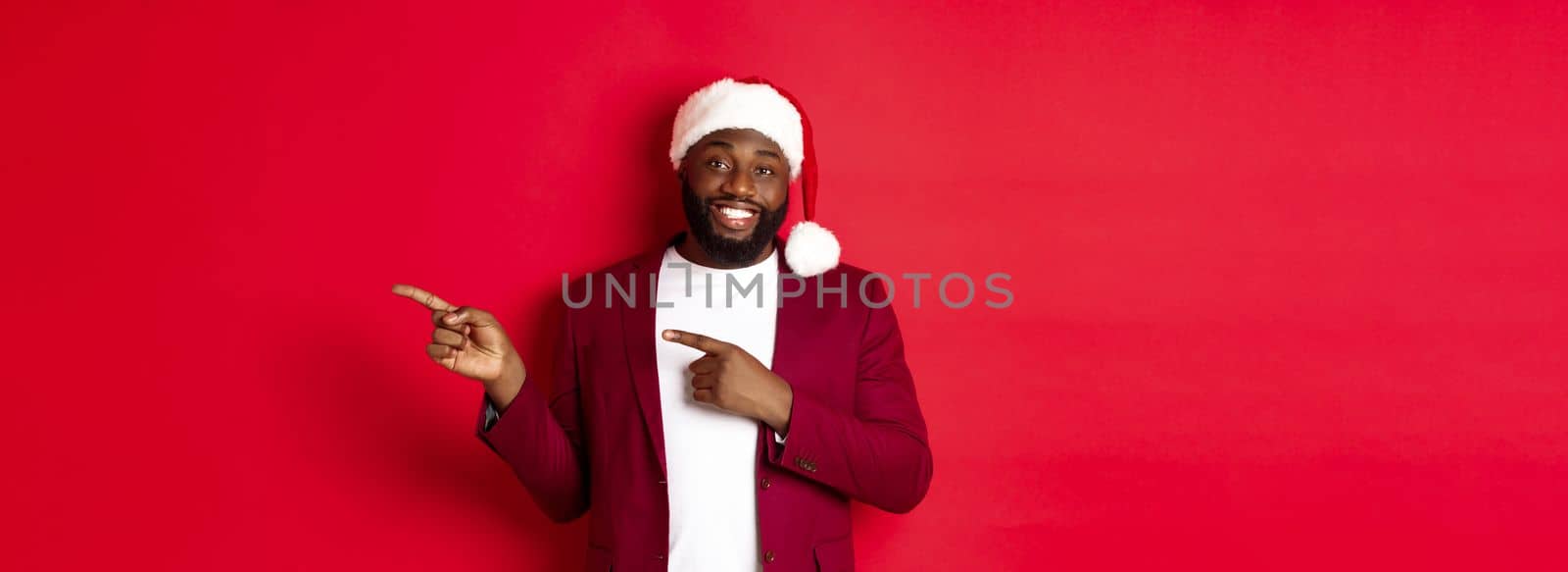 Christmas, party and holidays concept. Smiling Black man with beard and santa hat, pointing fingers right at showing logo, standing over red background.