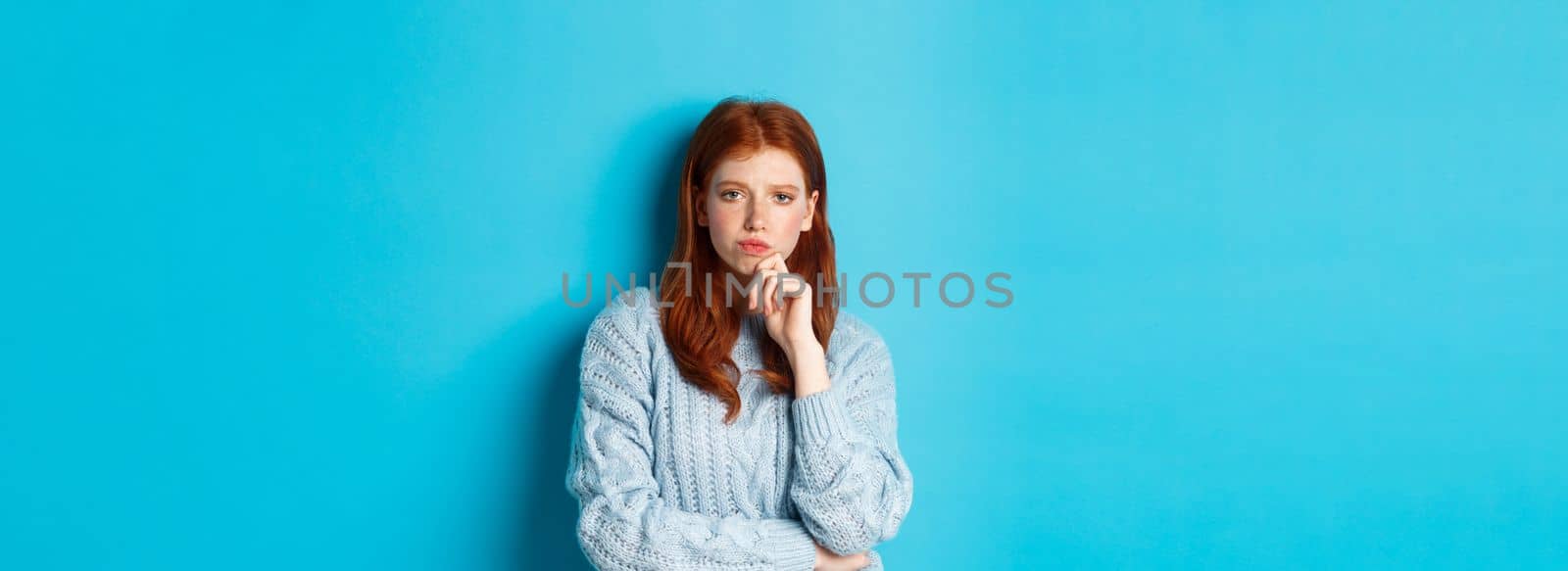 Thoughtful teenage redhead girl staring at camera, pondering ideas, making decision with serious face, standing over blue background.