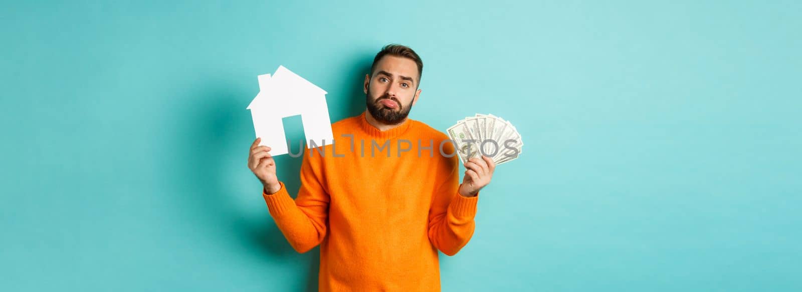 Real estate concept and mortgage concept. Sad and confused man showing money and paper house maket, shrugging upset, standing over light blue background.