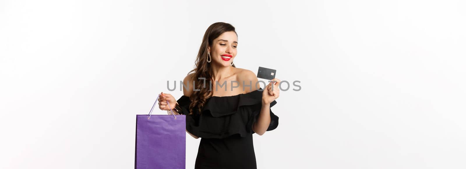 Stylish young woman in black dress going shopping, holding bag and credit card, smiling pleased, standing over white background.