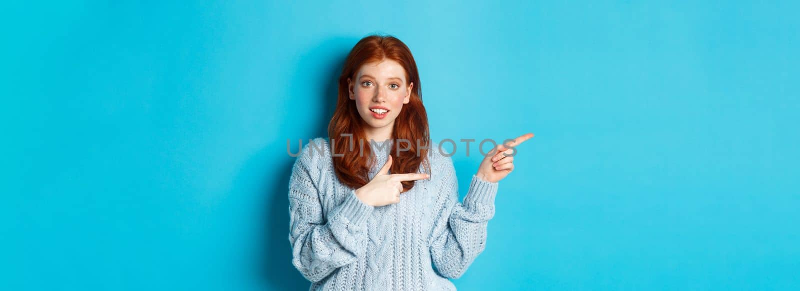 Image of pretty redhead girl in sweater, pointing fingers left at logo, smiling curious, standing over blue background.