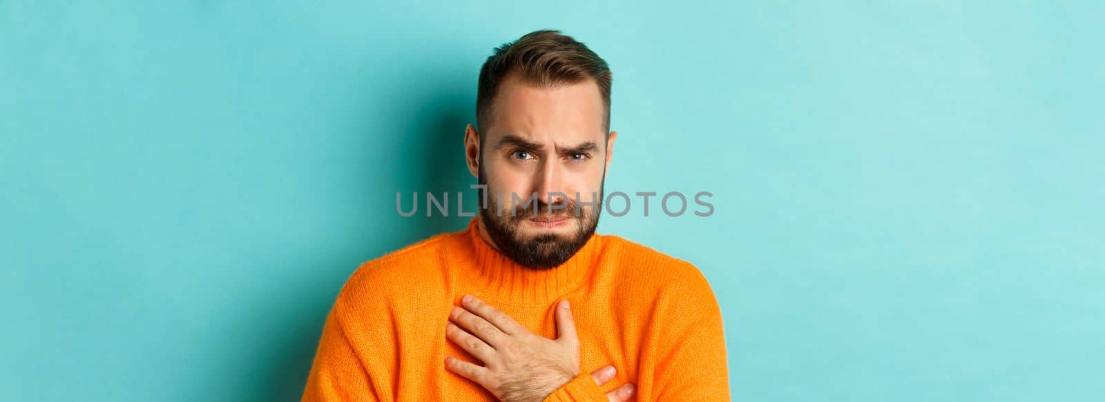 Close-up of shocked and startled man touching chesk, looking displeased and worried, standing against light blue background.