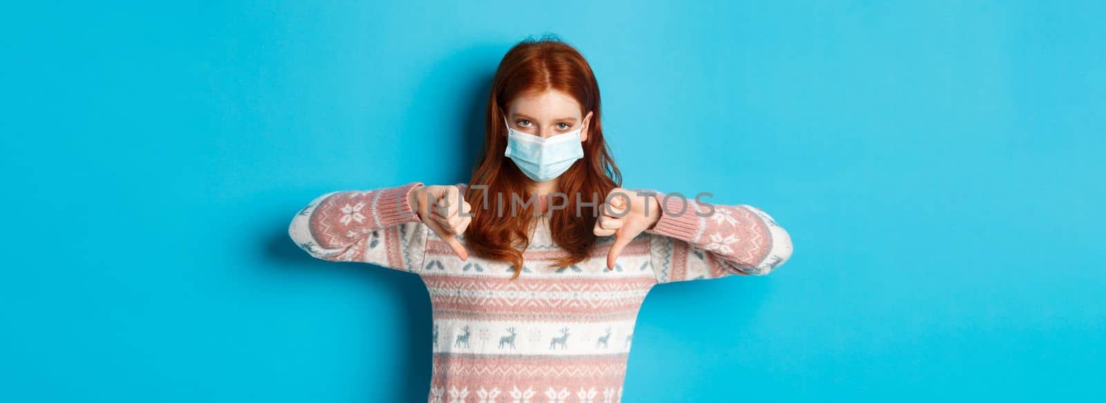 Winter, covid-19 and pandemic concept. Upset and angry redhead girl in face mask showing disapproval, thumbs down in dislike, standing over blue background.