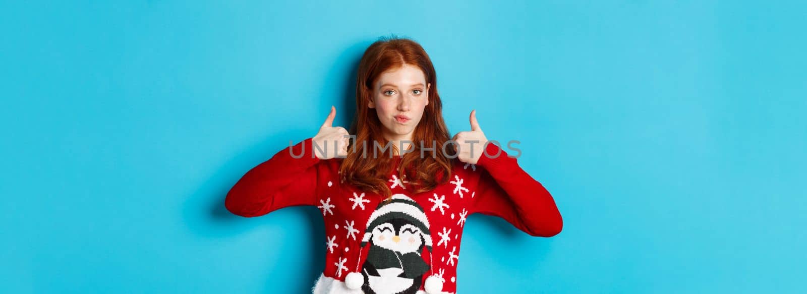 Winter holidays and Christmas Eve concept. Impressed redhead girl in xmas sweater, nod in approval and showing thumb up, praise good product, standing over blue background.
