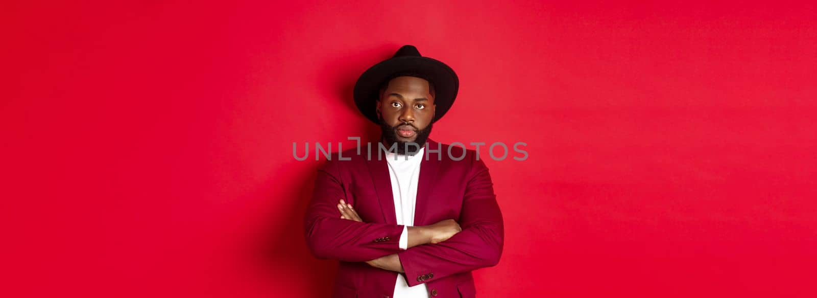 Serious black man standing in party outfit and waiting, cross arms on chest staring at camera, red background.