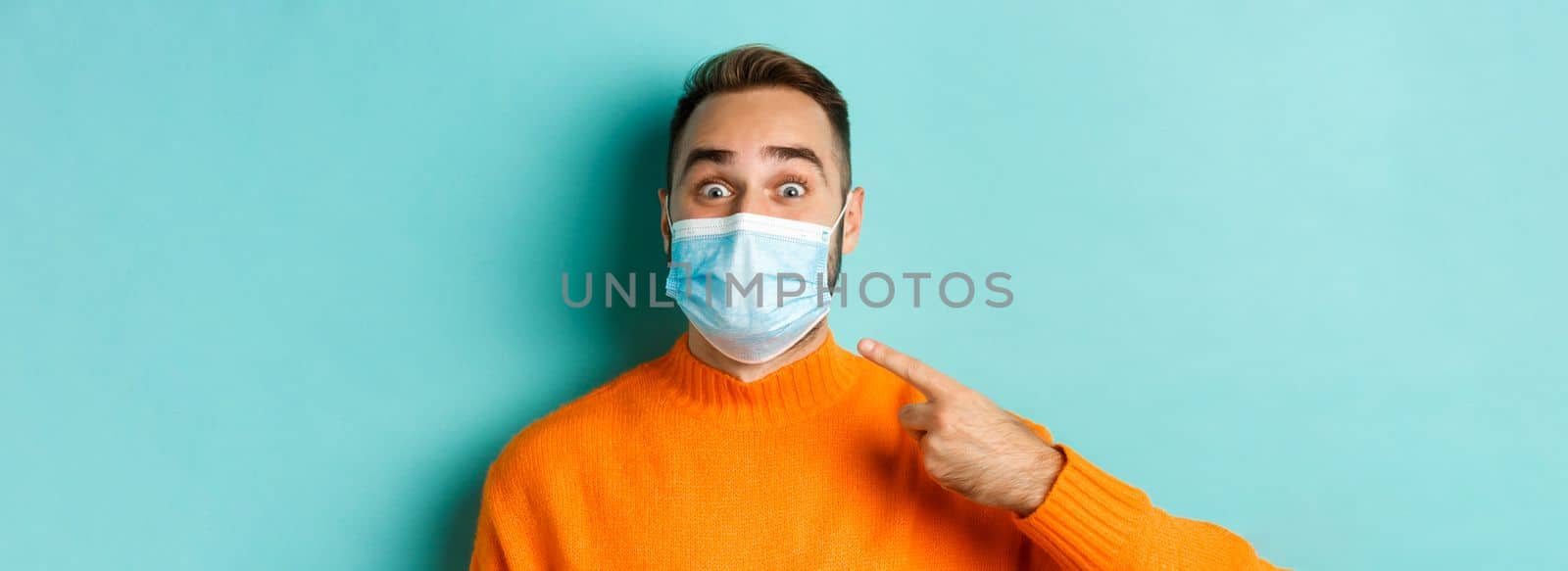 Covid-19, social distancing and quarantine concept. Close-up of cheerful man pointing finger at his medical mask, standing over turquoise background.