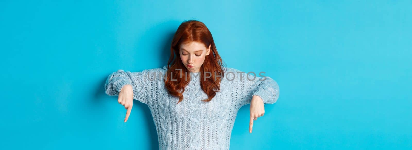 Winter holidays and people concept. Intrigued redhead girl, pointing and looking down thoughtful, making choice, standing over blue background.