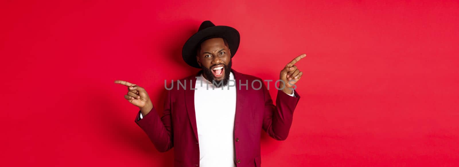Winter holidays and shopping concept. Cheerful Black man smiling and showing two promo, pointing fingers sideways at copy spaces, red background.