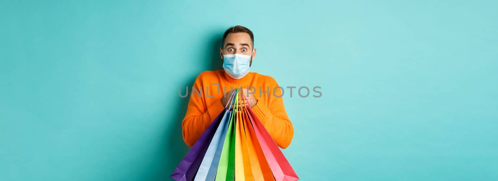 Covid-19, pandemic and lifestyle concept. Excited man in face mask showing shopping bags and rejoicing from discounts, standing over turquoise background.
