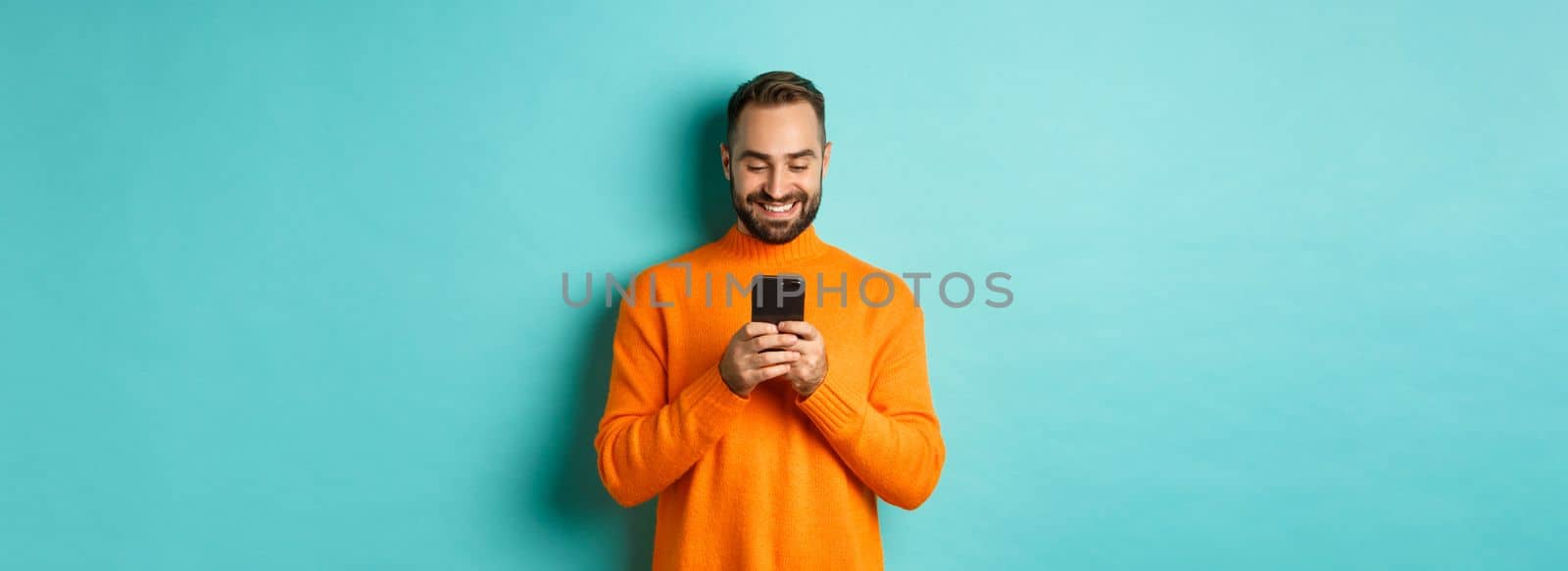 Handsome man smiling and texting message on mobile phone, communicating online, standing over light blue background.