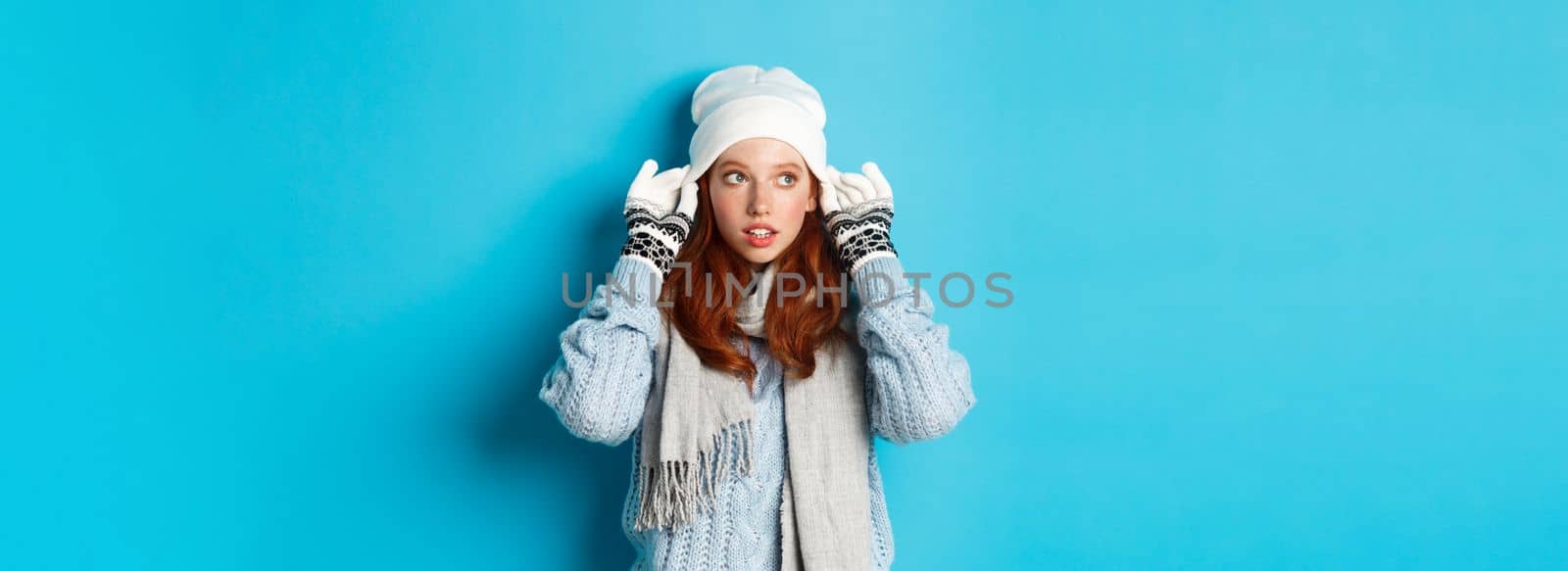 Winter and holidays concept. Cute redhead girl going outside, put on beanie and gloves, looking left, standing over blue background.