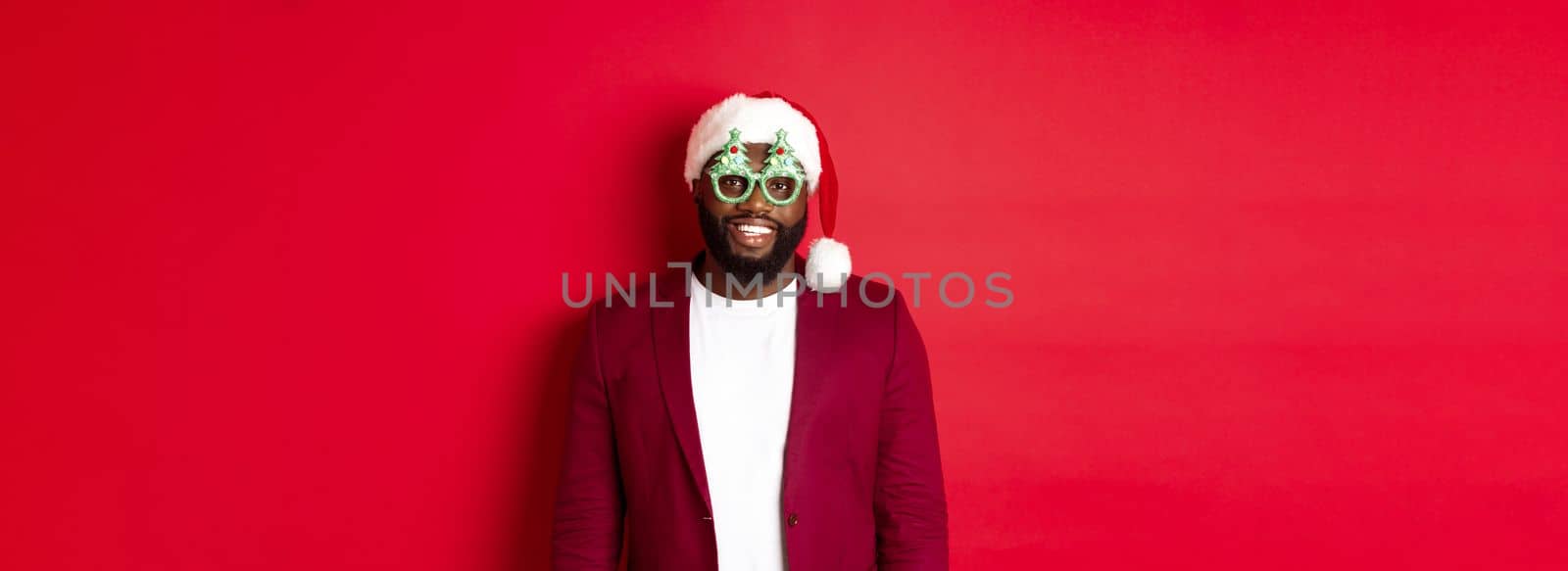 Funny Black man in santa hat and party glasses celebrating Christmas, smiling happy and wishing merry xmas, standing over red background.