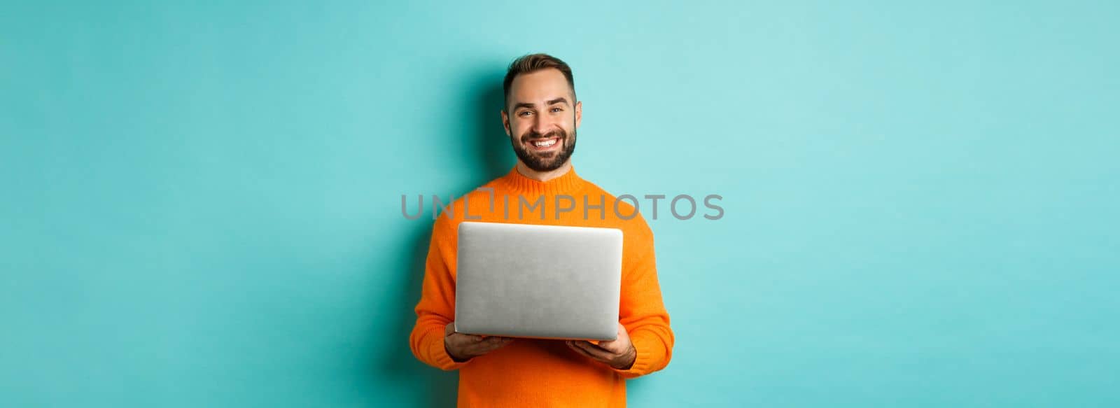 Handsome freelancer working on laptop and smiling, standing in orange sweater over light blue background.