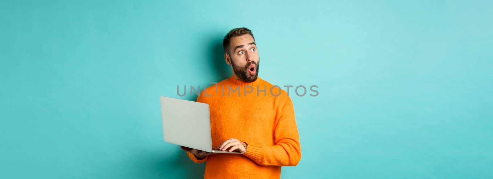 Handsome man doing online shopping, looking up thoughtful while using laptop, standing over light blue background by Benzoix