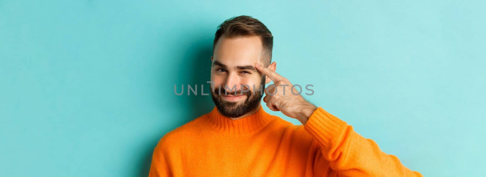 Image of young man in orange sweater, smiling and pointing at head, having an idea, praising good thought, standing over light blue background.