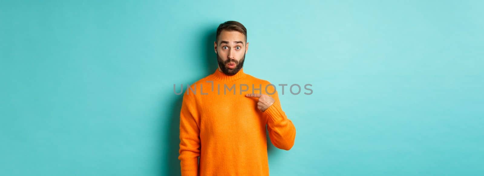 Man pointing at himself with surprise face, being chosen, standing confused against light blue background.