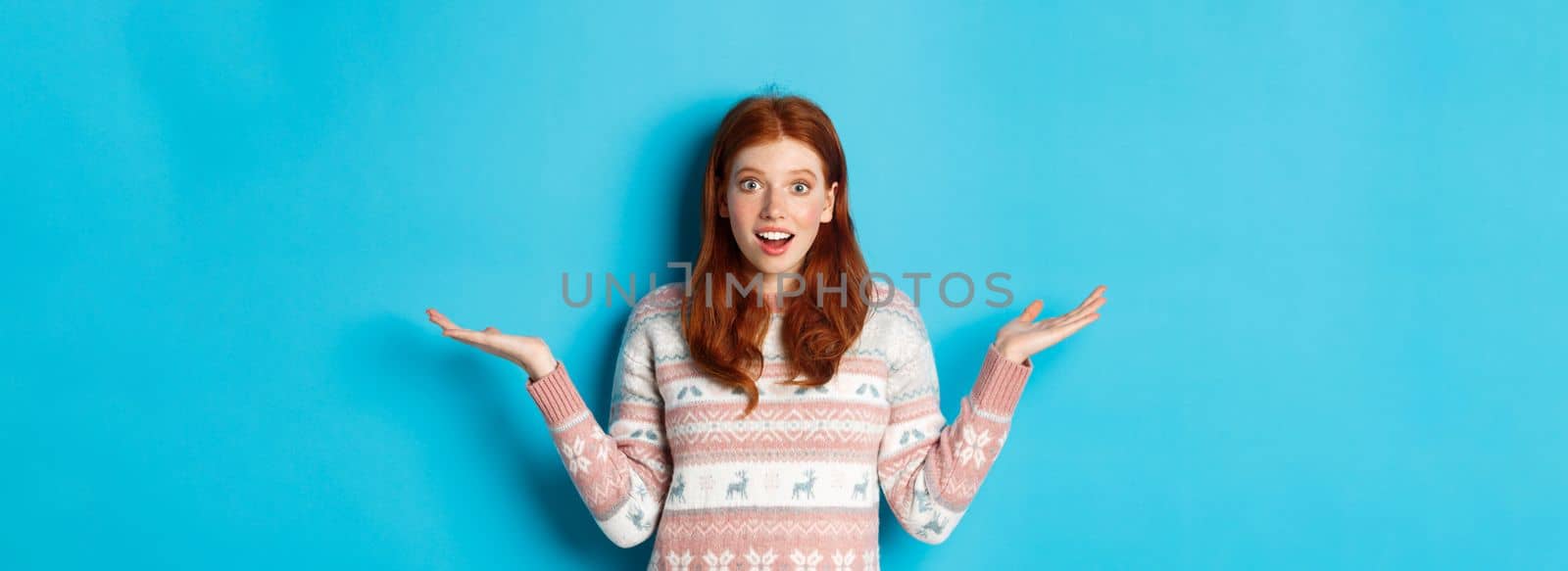 Image of excited redhead girl react to good news, looking surprised, spread hands sideways and smiling, standing in winter sweater against blue background.