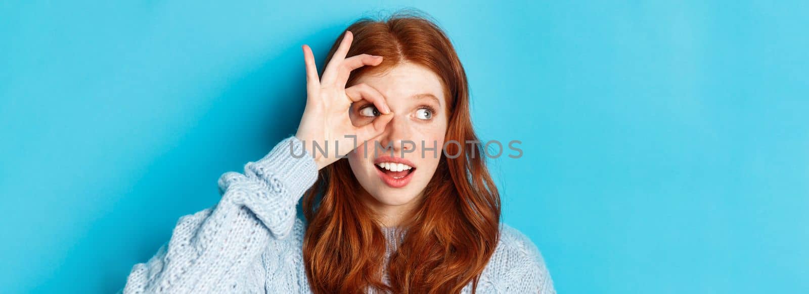 Headshot of pretty redhead girl in sweater, looking left at promo with okay sign over eye, standing against blue background.