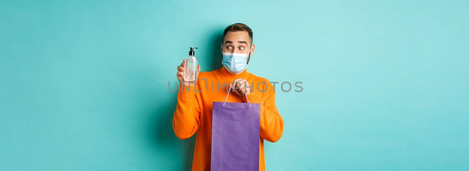 Coronavirus, pandemic and lifestyle concept. Man in face mask showing shopping bag and hand sanitizer, standing over turquoise background.